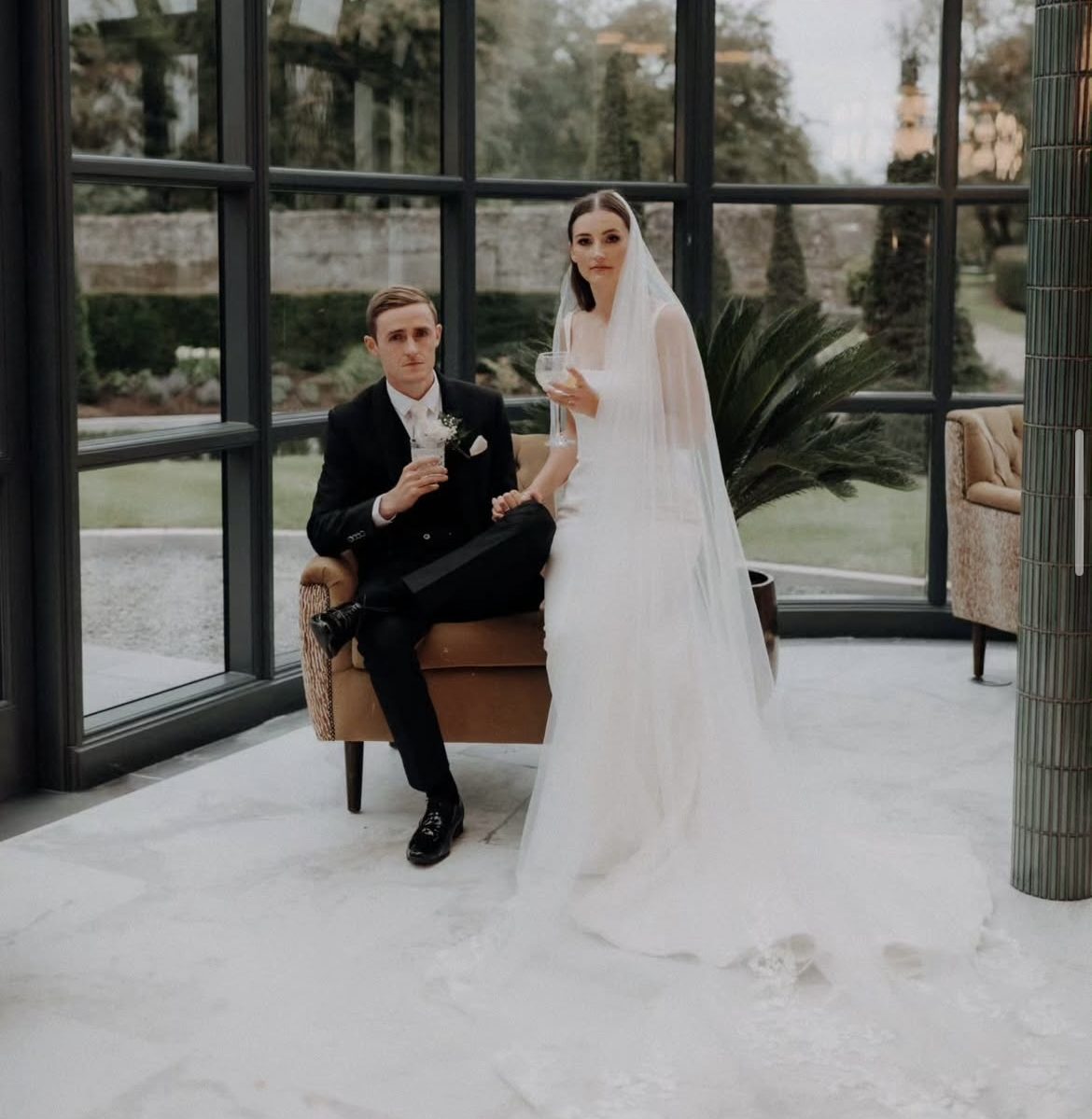 Groom in a black suit seated beside his bride in a glass conservatory
