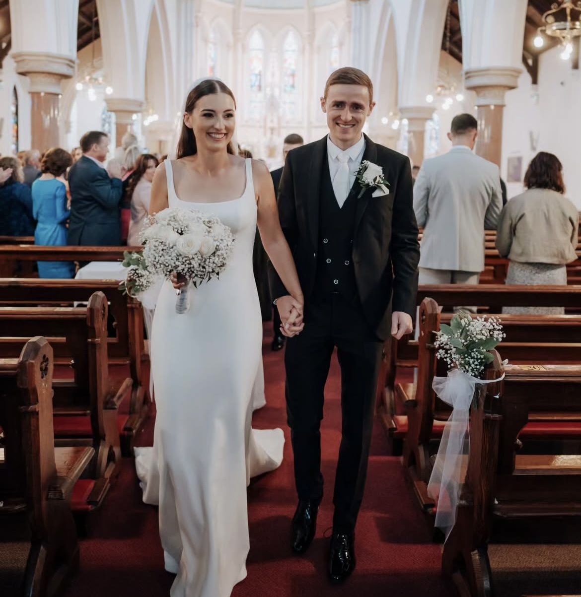 Groom in a black three-piece suit walking down the church aisle with his bride