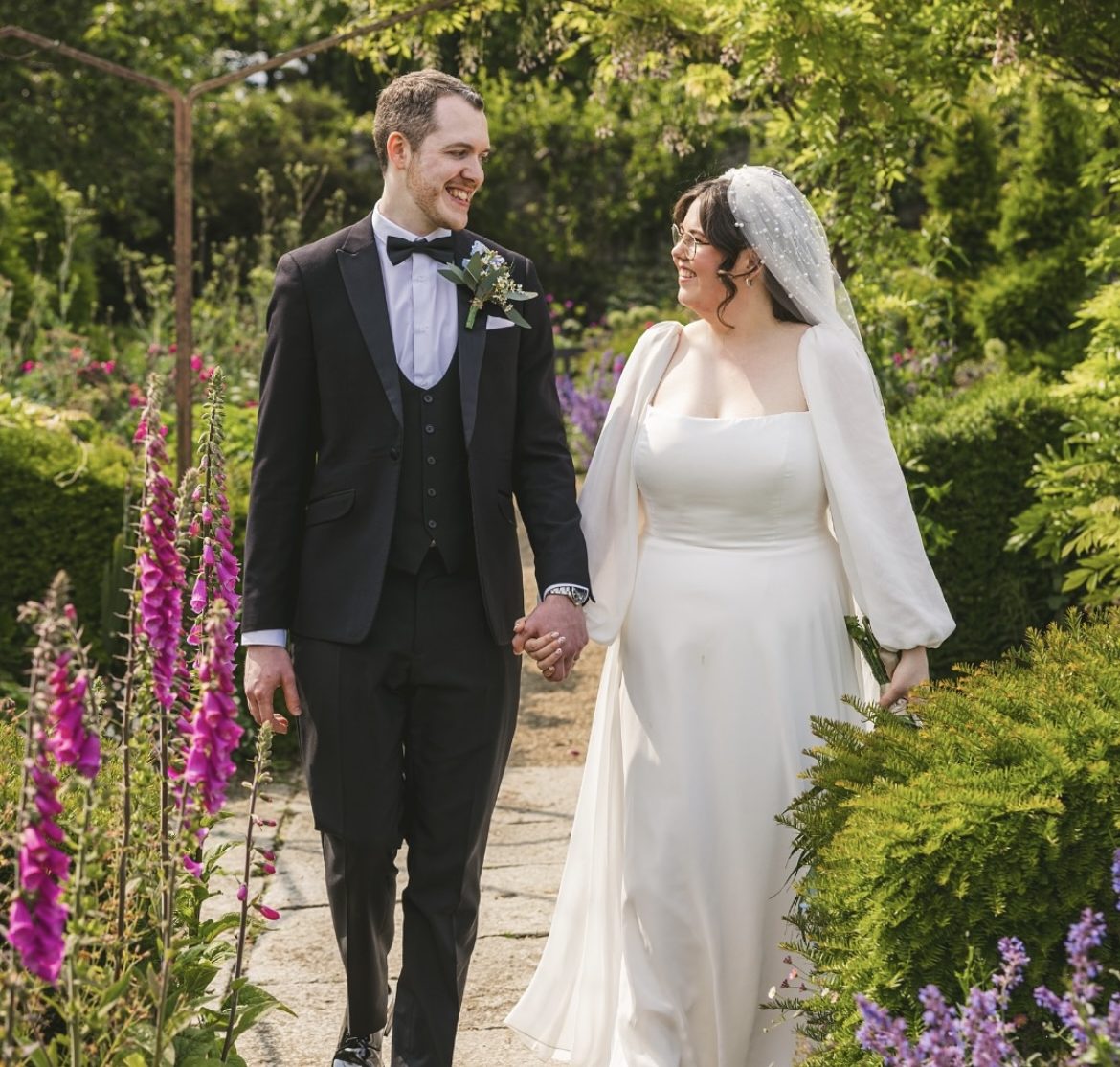 Groom in a black three-piece suit walking with his bride through a garden